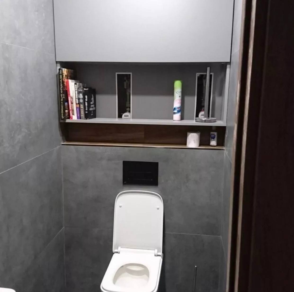 Nowoczesna łazienka z szarymi płytkami, podwieszaną toaletą z deską, wnęką z półkami na książki i dekoracje oraz czarnym przyciskiem spłukującym.