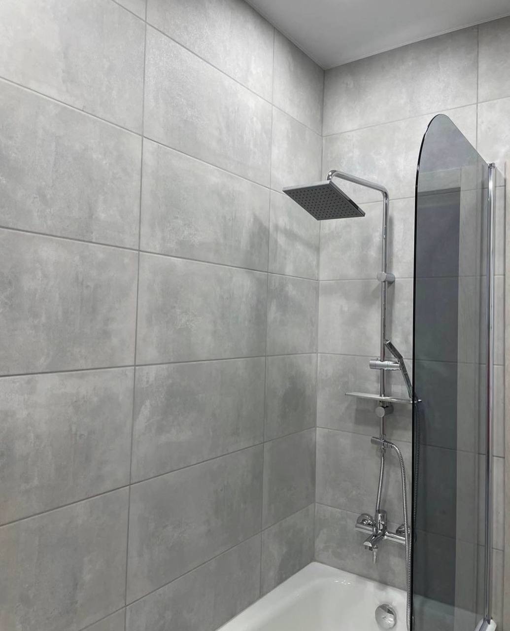 Minimalistyczna łazienka z szarymi, matowymi płytkami imitującymi beton, kwadratowa deszczownica i szklany parawan nawannowy.