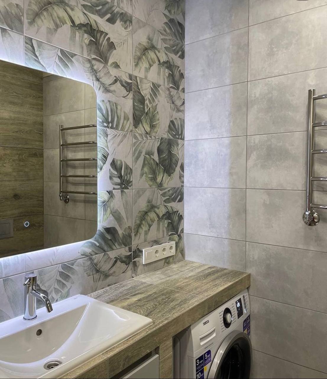 Umywalka w łazience z płytkami w motywy roślinne, drewnopodobny blat i pralka pod blatem.