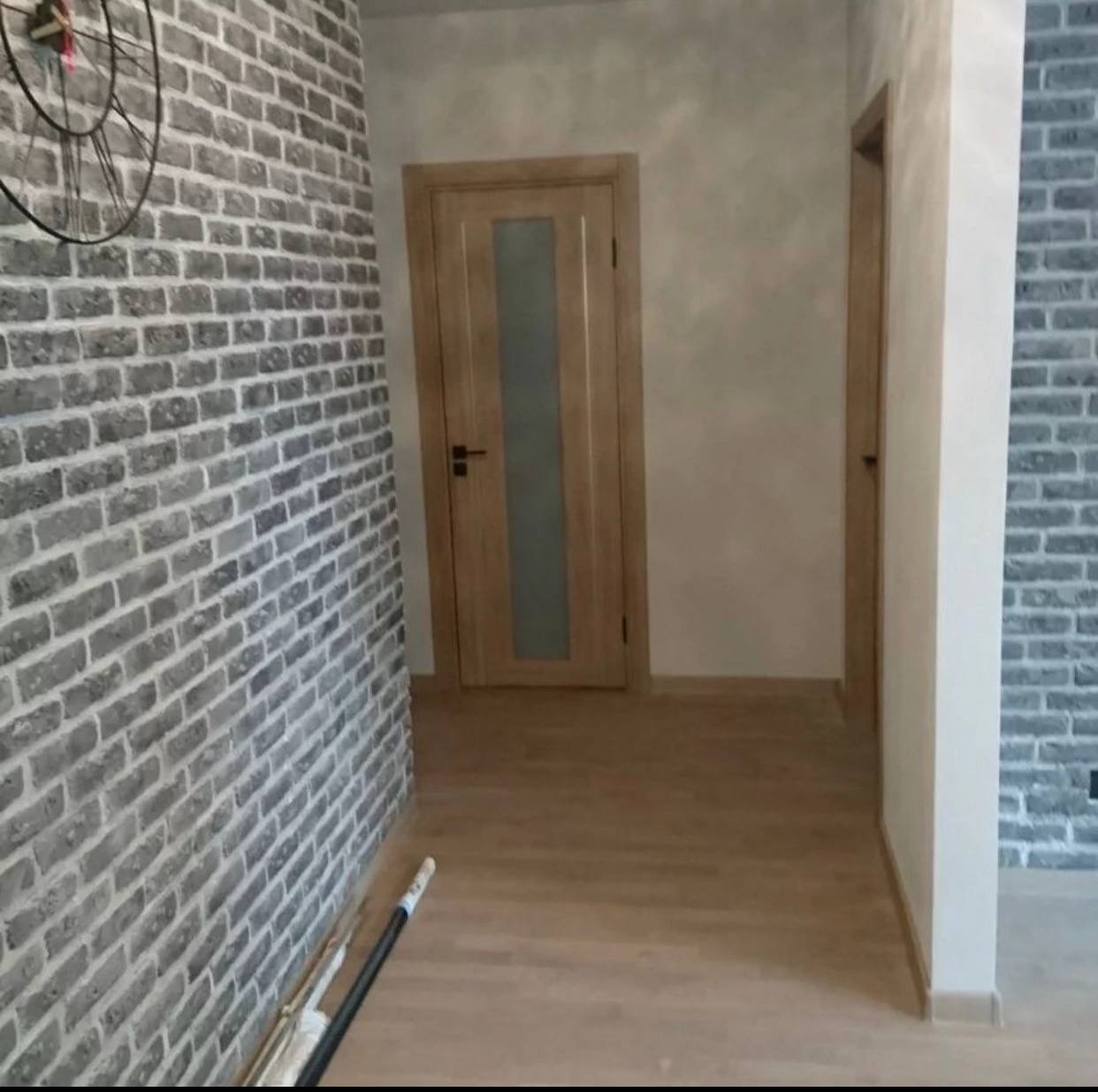 Korytarz z dekoracyjną ścianą z szarej cegły, dwoje drewnianych drzwi i podłoga z paneli, widoczne narzędzia przy ścianie.