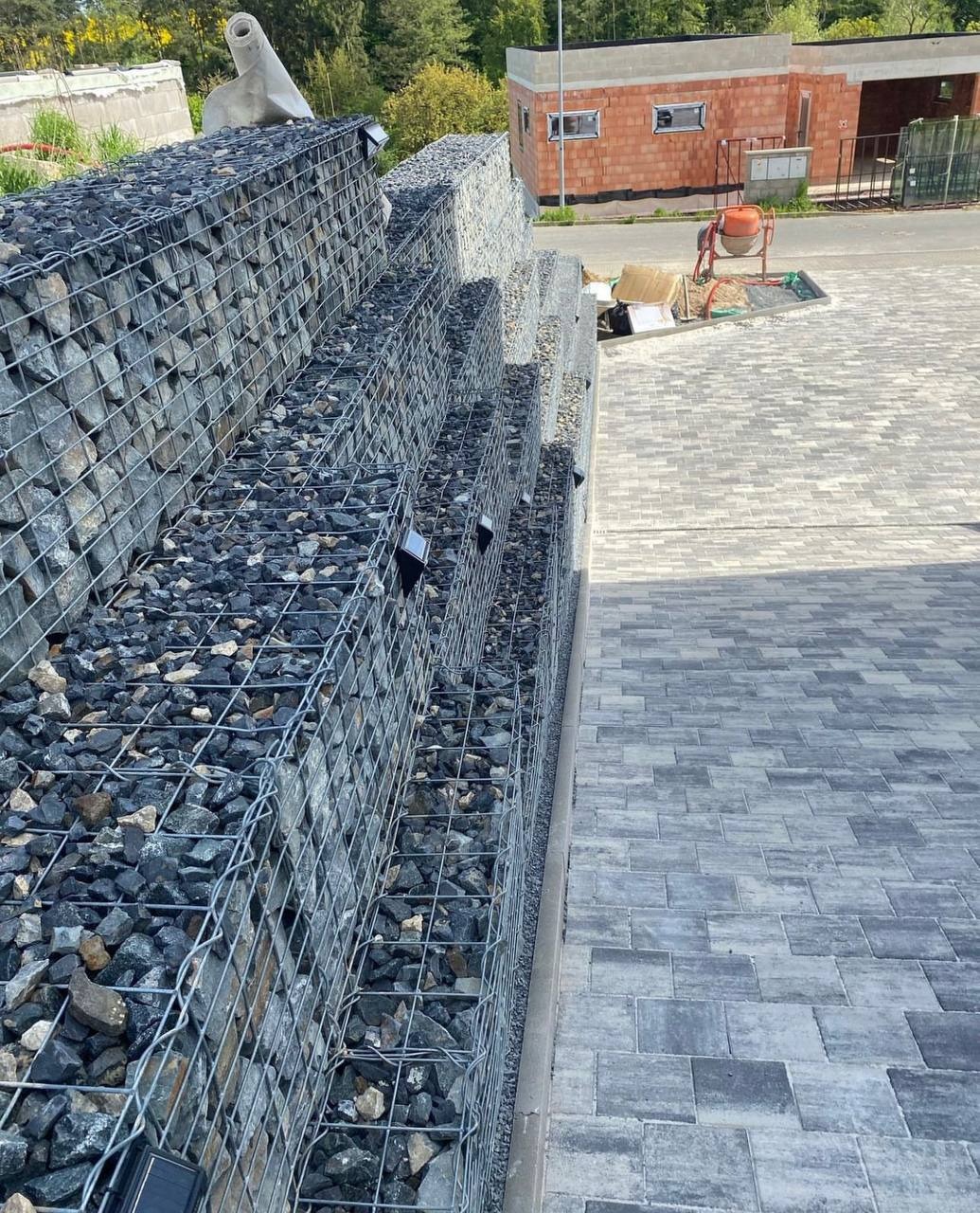 Gabionowa ściana oporowa z czarnym grysem, ułożona schodkowo, obok szara kostka brukowa, w tle budynek w stanie surowym otwartym i betoniarka.