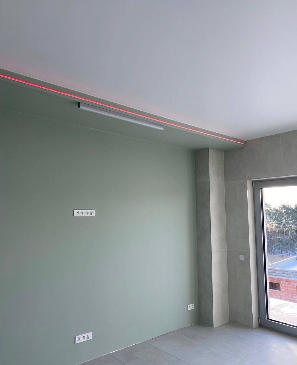 Świeżo wykończone wnętrze z zieloną ścianą, szarą podłogą i oświetleniem LED pod sufitem, widoczne gniazdka elektryczne i okno z widokiem na zewnątrz.