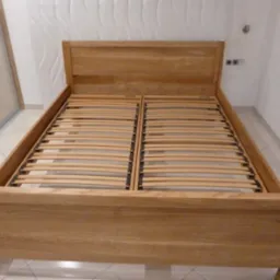 Rama łóżka z jasnego drewna z listwami, widok z przodu, w tle ściana z dekoracyjną fakturą i szafa przesuwna.