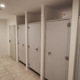 Firma Remontowo-Budowlana Jacoob Jakub Jankowski - Publiczna toaleta z kabinami w odcieniach szarości i beżu, widoczne umywalki. Minimalistyczny design, płytki na podłodze i ścianach, oświetlenie sufitowe.