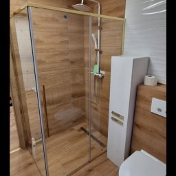 Firma Remontowo-Budowlana Jacoob Jakub Jankowski - Nowoczesna kabina prysznicowa z złotymi profilami, drewnopodobne płytki na ścianach i podłodze, biała szafka łazienkowa i toaleta. Minimalistyczny design.