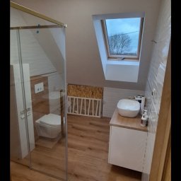 Firma Remontowo-Budowlana Jacoob Jakub Jankowski - Jasna łazienka na poddaszu z kabiną prysznicową, umywalką na szafce i oknem dachowym. Drewnopodobne płytki na podłodze i białe płytki na ścianach.