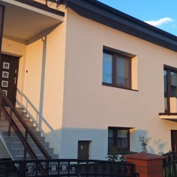 Firma Remontowo-Budowlana Jacoob Jakub Jankowski - Dwupiętrowy dom z beżową elewacją w ciepłym świetle popołudniowym. Widoczne schody wejściowe, balkon i okna z brązowymi ramami. Ciemny dach i ogrodzenie.