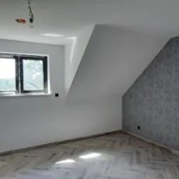 Pokój na poddaszu po remoncie: białe ściany, szara ściana z fakturą, parkiet ułożony w jodełkę, czarna rama okienna, wisząca żarówka.