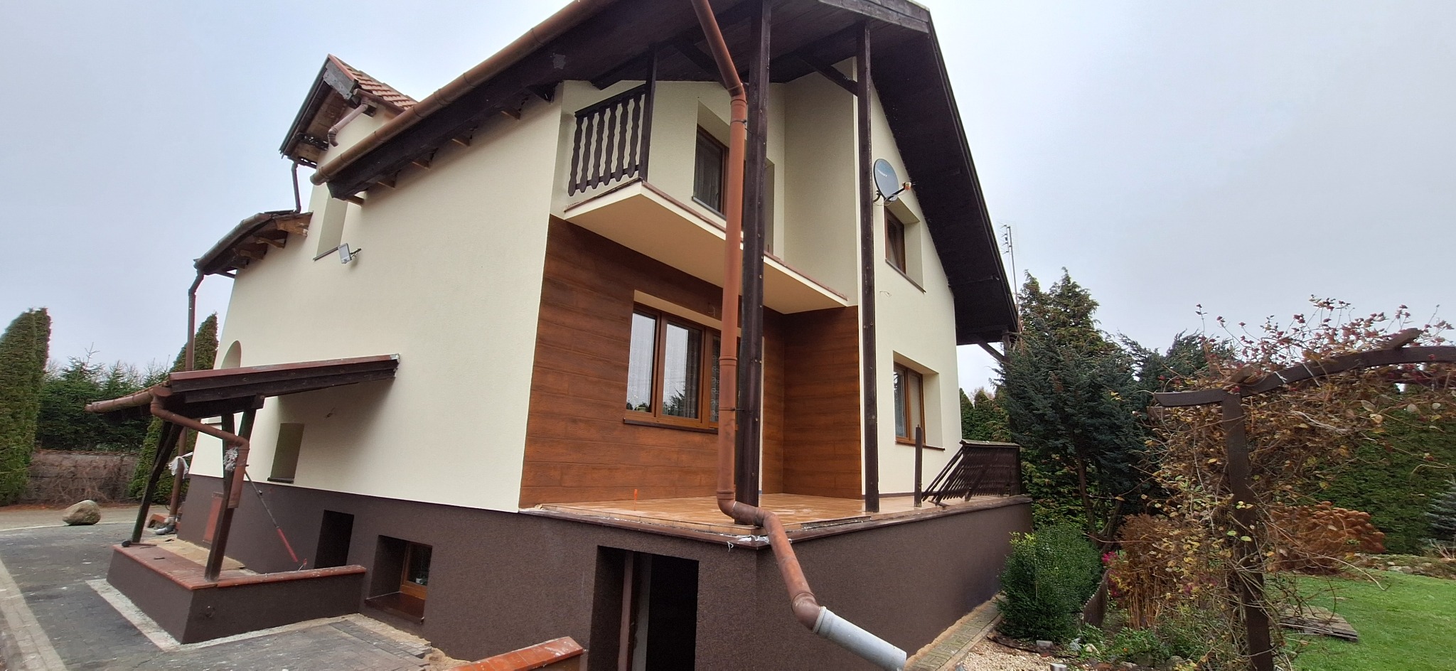 Dwukondygnacyjny dom z beżową elewacją i brązowymi akcentami, w tym drewnianą okładziną i rynnami. Widoczny balkon i satelita na ścianie. Ujęcie z poziomu gruntu.