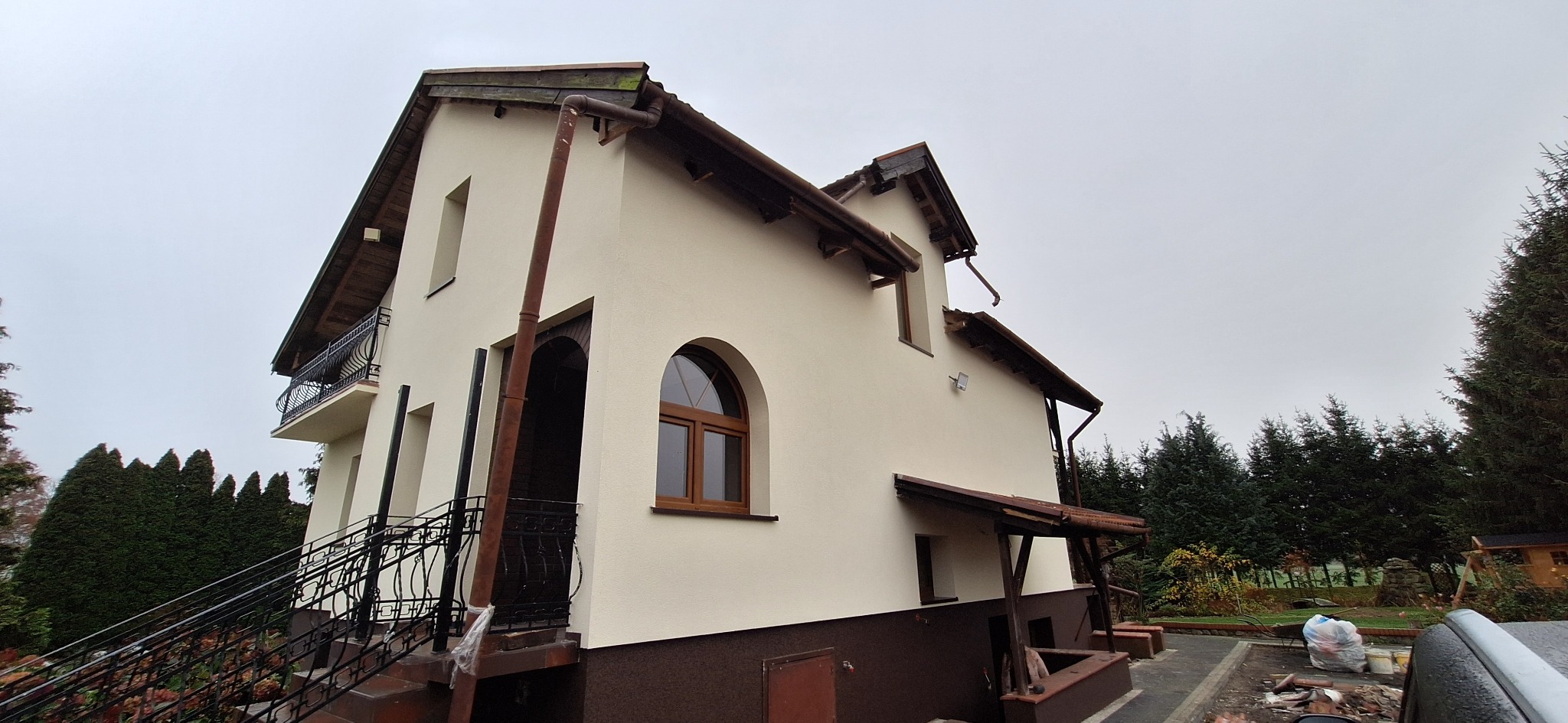 Dwukondygnacyjny dom z beżową elewacją i brązowymi akcentami, widoczny z dołu pod kątem. Na pierwszym planie schody z czarną, ozdobną balustradą.