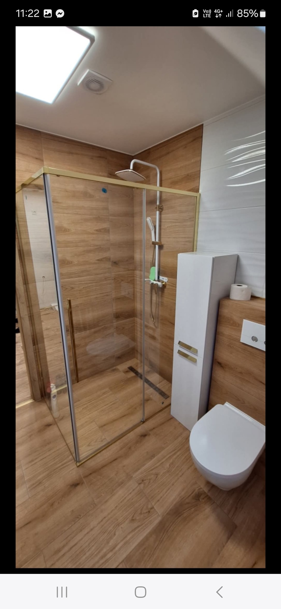 Nowoczesna kabina prysznicowa z złotymi profilami, drewnopodobne płytki na ścianach i podłodze, biała szafka łazienkowa i toaleta. Minimalistyczny design.