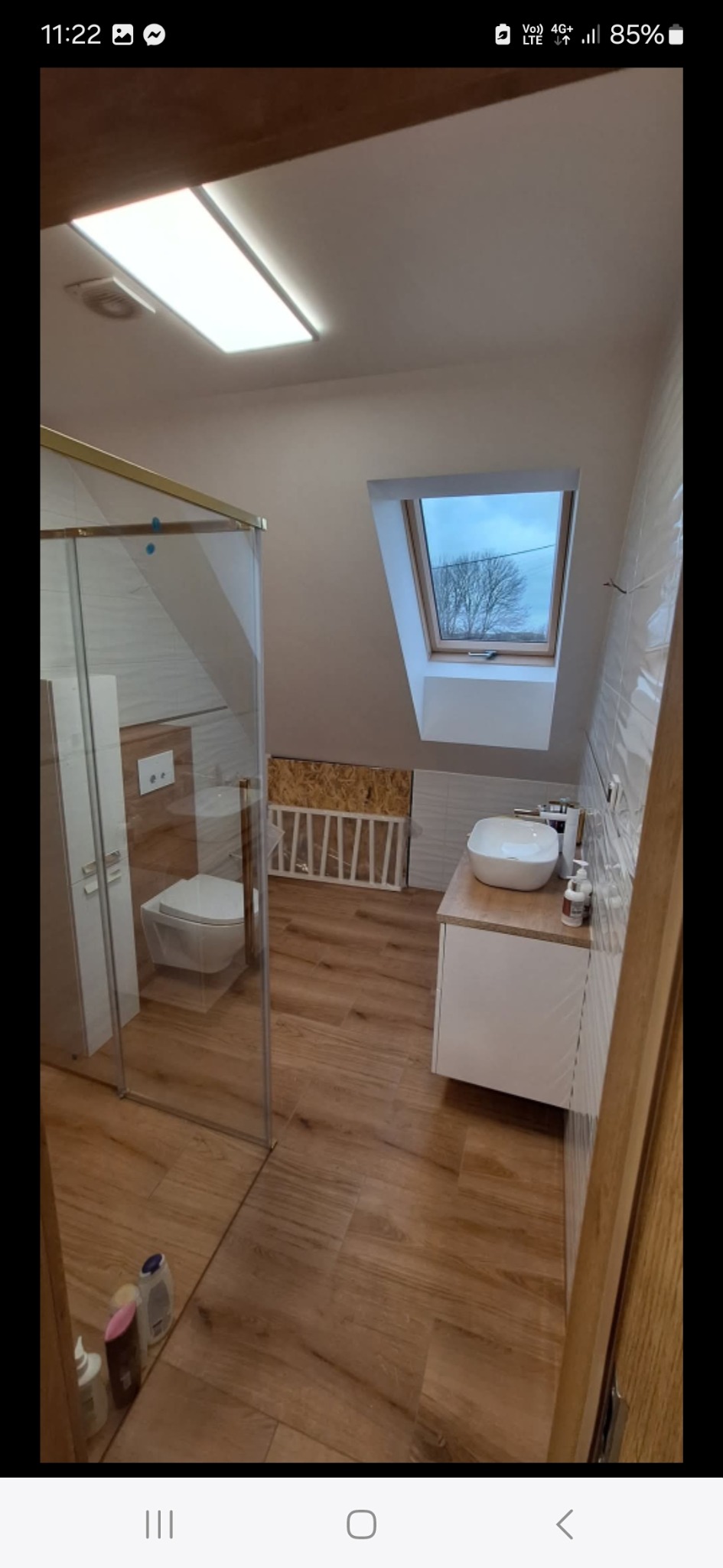 Jasna łazienka na poddaszu z kabiną prysznicową, umywalką na szafce i oknem dachowym. Drewnopodobne płytki na podłodze i białe płytki na ścianach.