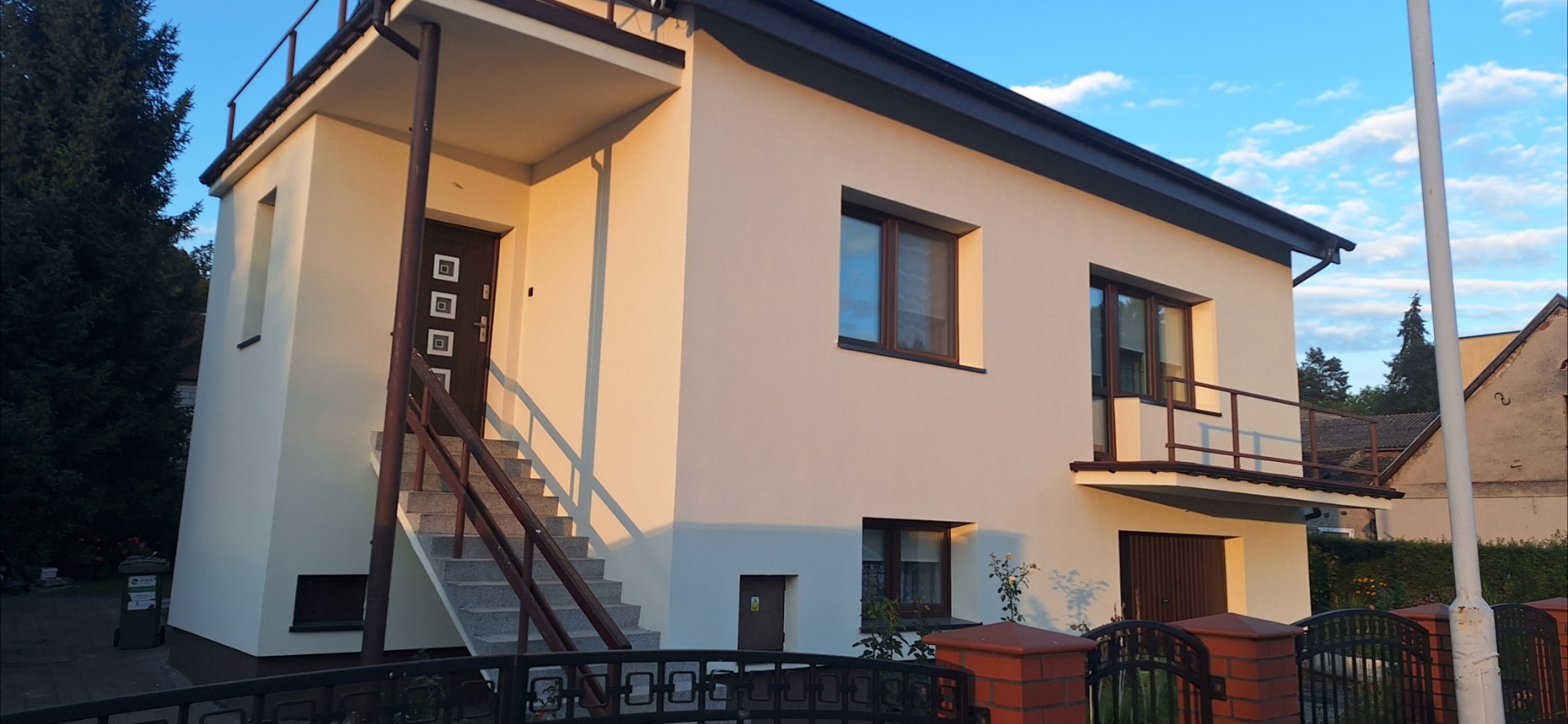 Dwupiętrowy dom z beżową elewacją w ciepłym świetle popołudniowym. Widoczne schody wejściowe, balkon i okna z brązowymi ramami. Ciemny dach i ogrodzenie.