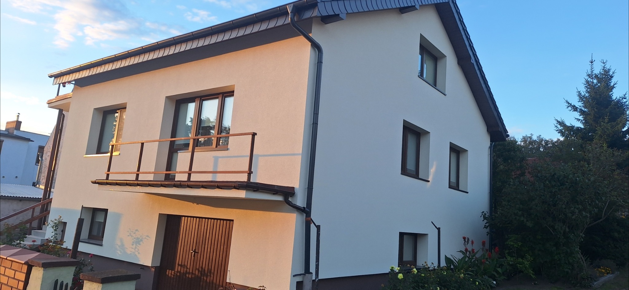 Dwupiętrowy dom z balkonem po renowacji elewacji, w ciepłym świetle zachodzącego słońca. Widoczne okna z brązowymi ramami i ciemny dach.
