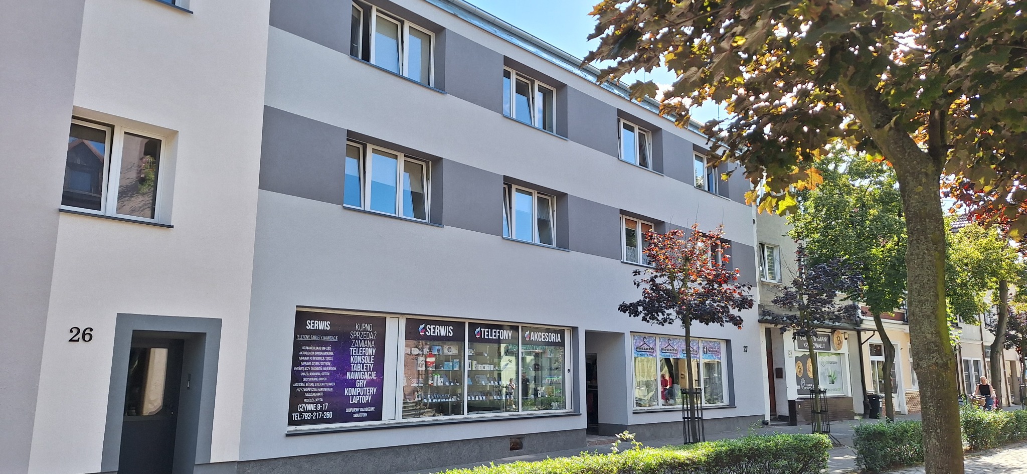 Odnowiony budynek z szarą elewacją i białymi oknami w słoneczny dzień. Widoczne drzewa i krzewy przed budynkiem. Numer 26 nad wejściem.