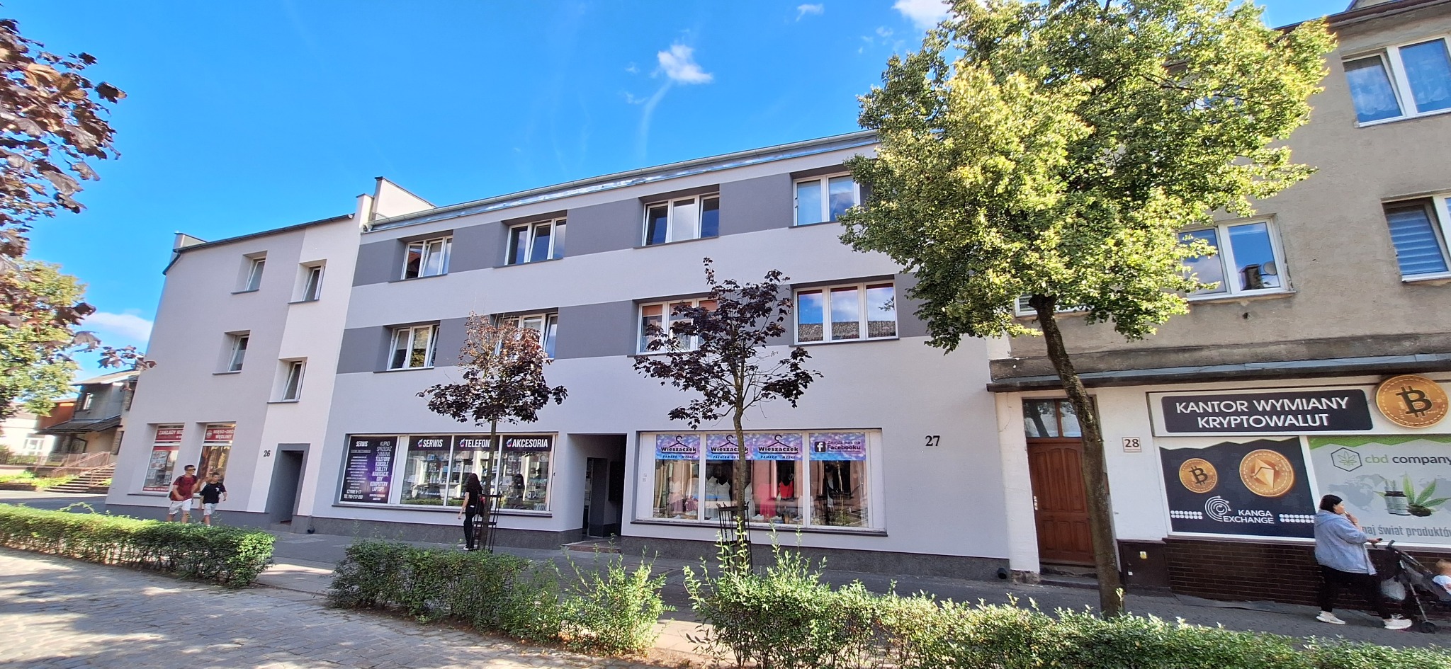 Odnowiona elewacja budynku z szarymi pasami, widok z poziomu ulicy, zieleń miejska, błękitne niebo. Budynek z numerami 26, 27, 28.
