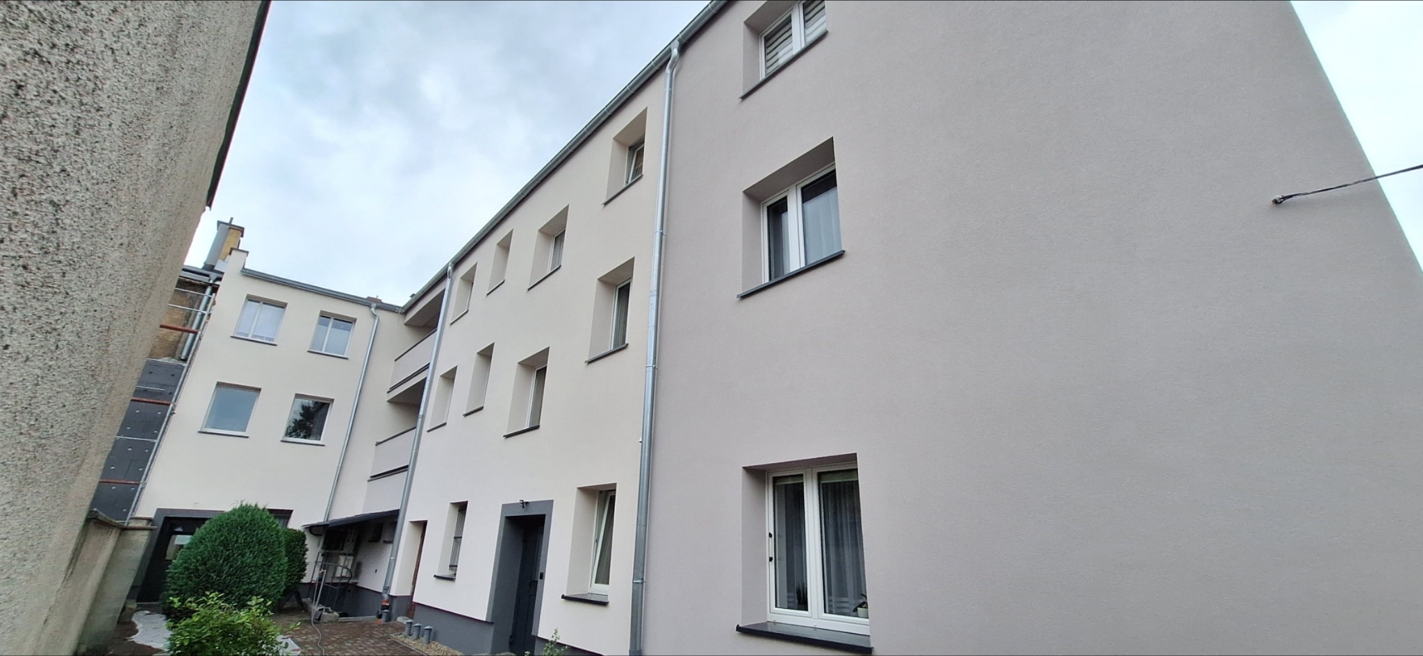 Odnowiona elewacja budynku wielorodzinnego w jasnym odcieniu beżu, z widocznymi oknami i rynnami. Fragment ocieplenia na sąsiednim budynku. Ujęcie z dołu.