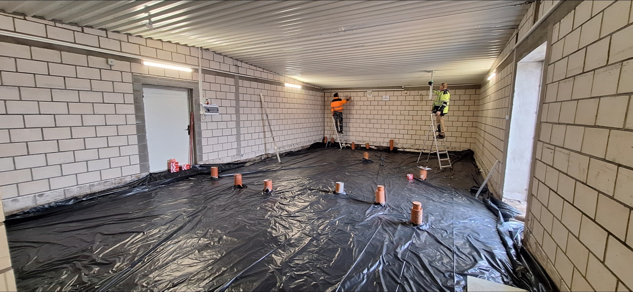 Wnętrze w trakcie prac: folia na podłodze, wystające rury, dwóch pracowników na drabinach przy ścianach z białej cegły. Widoczne oświetlenie i surowy strop.