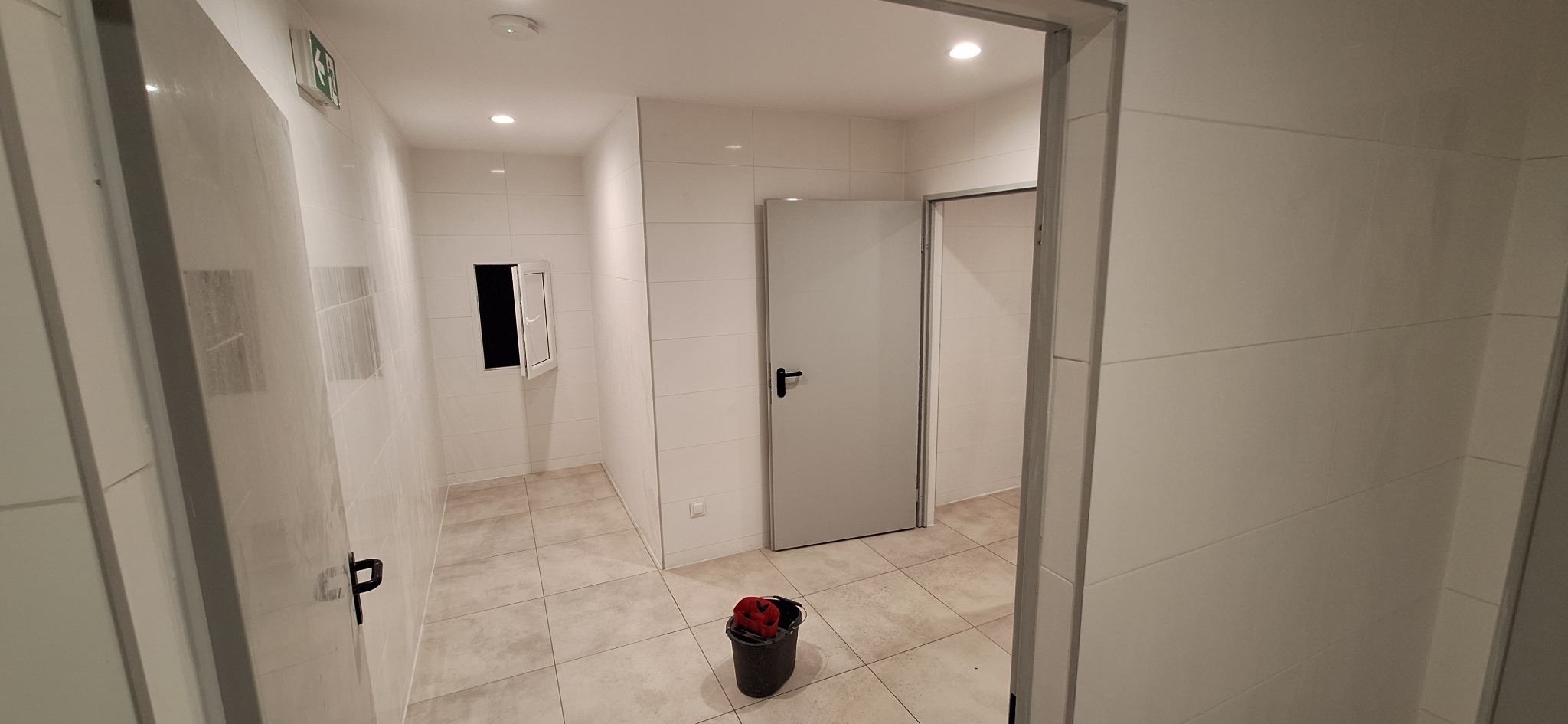 Wykończona łazienka w jasnych płytkach z otwartym oknem i drzwiami. Na podłodze stoi wiadro z narzędziami. Widok z korytarza.