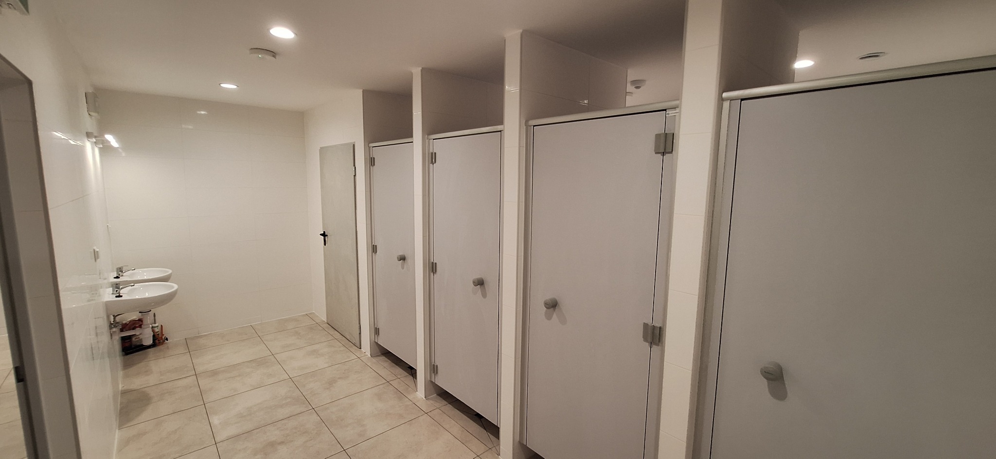 Wnętrze publicznej toalety z kabinami i umywalkami. Jasne płytki na ścianach i podłodze. Widoczne detale wykończenia, oświetlenie sufitowe. Minimalistyczny design.