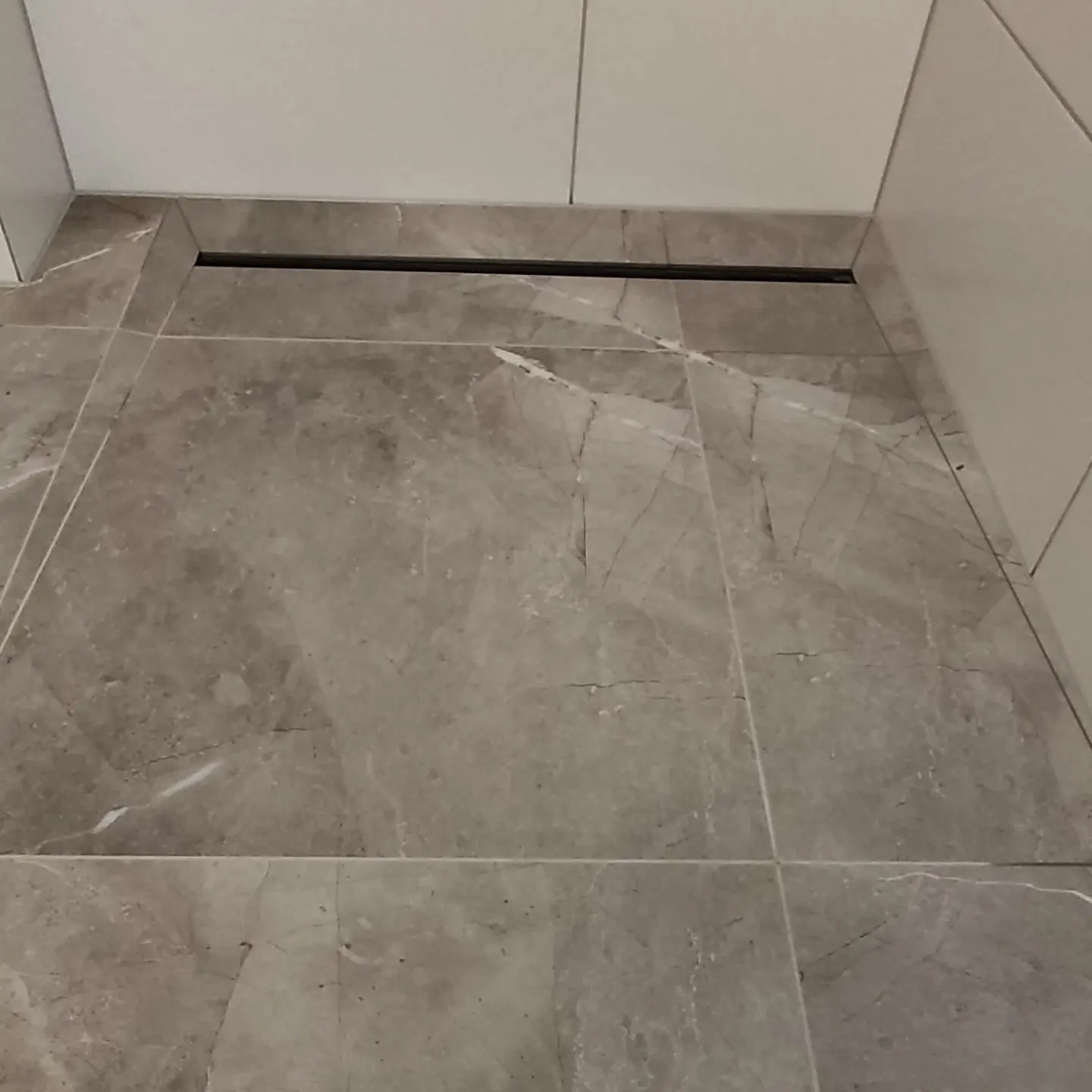 Szare marmurowe płytki podłogowe z czarnym odpływem liniowym przy ścianie w łazience.