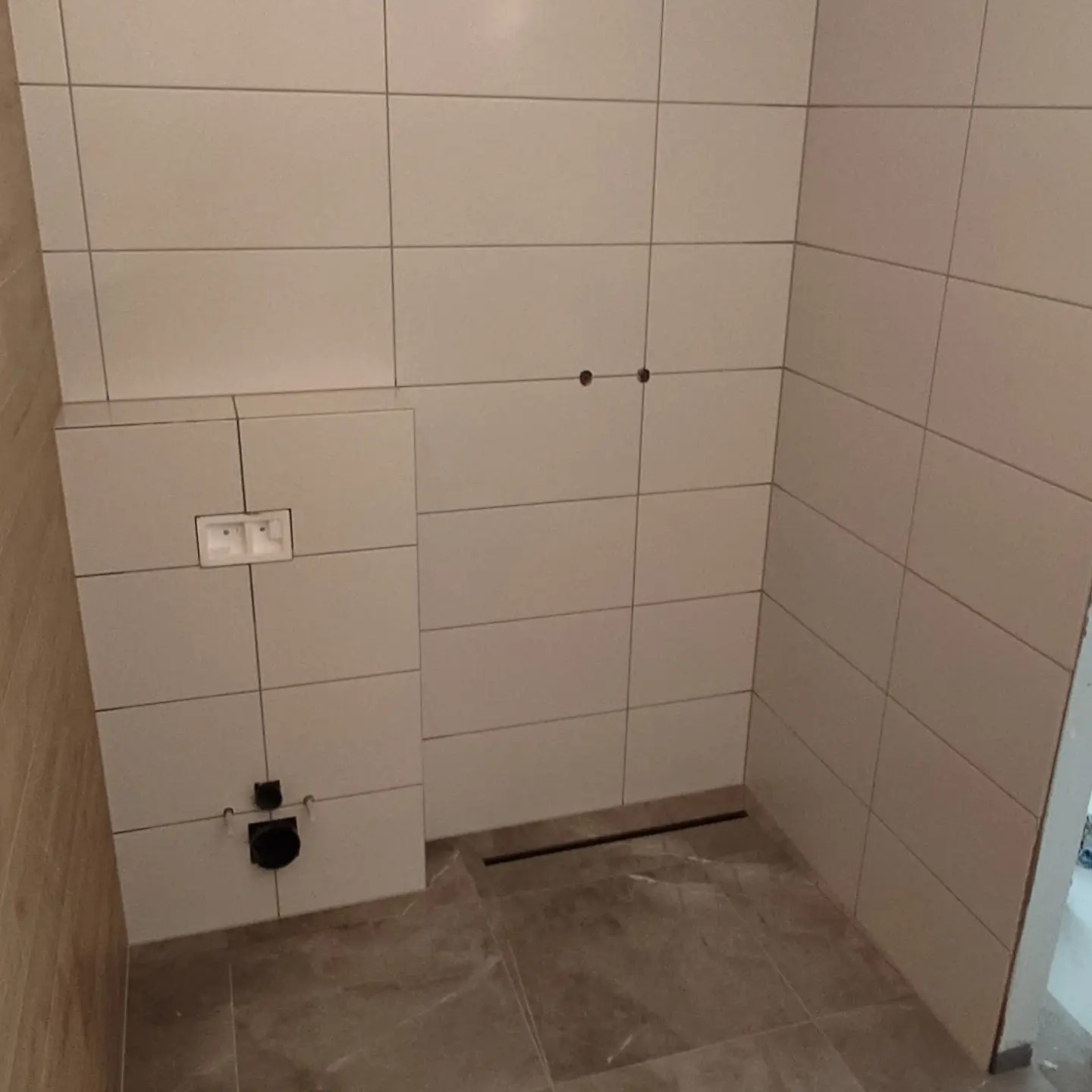 Wykończona łazienka z beżowymi płytkami na ścianach i podłodze, widoczny podtynkowy system spłukiwania, odpływ liniowy w podłodze i przygotowane otwory na baterię.