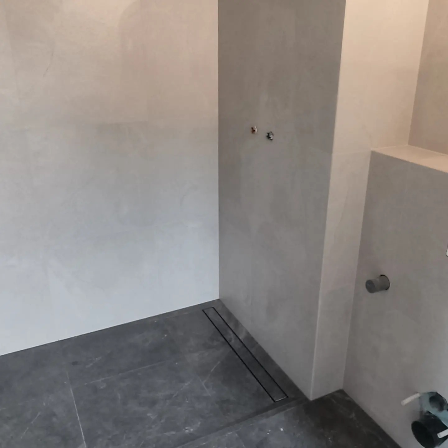 Surowa łazienka w trakcie remontu: szare płytki podłogowe, odpływ liniowy, przygotowane przyłącza wodne na ścianie, surowe ściany przygotowane do wykończenia.