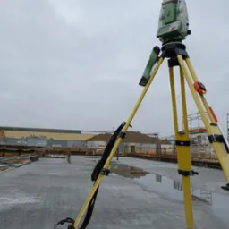 Zielono-szary teodolit na żółtym statywie ustawiony na mokrym, betonowym fundamencie budowlanym, w tle widoczne rusztowania i materiały budowlane pod pochmurnym niebem.