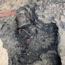 hidroallpl - Odkopana, skorodowana rura w ziemi z fragmentami cegieł i gruzu, widoczny fragment metalowego drutu.