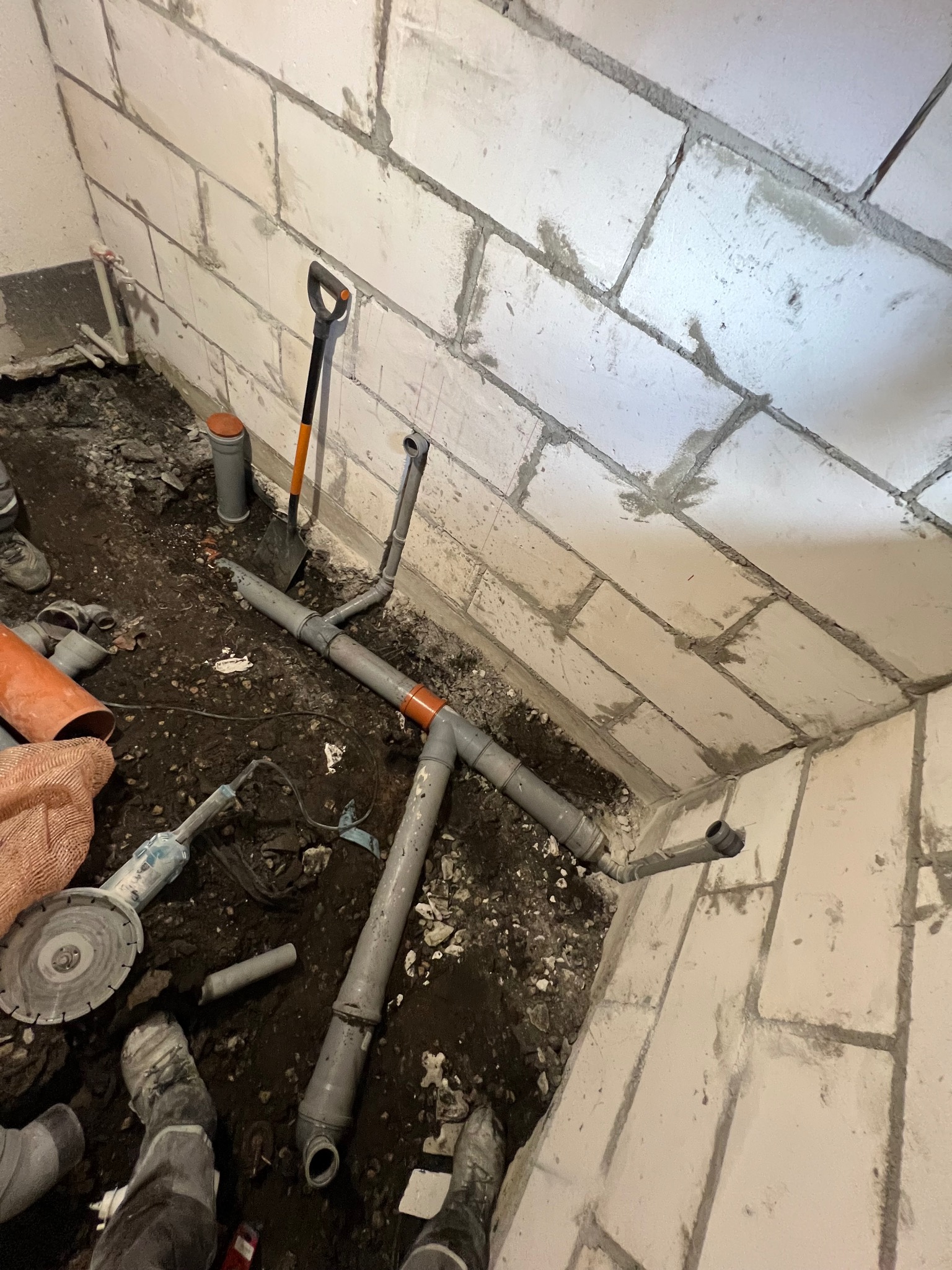 Wykop pod posadzkę z ułożonymi szarymi rurami kanalizacyjnymi, widoczne także narzędzia: szpachelka, kielnia i szlifierka kątowa, ściany z bloczków betonowych.