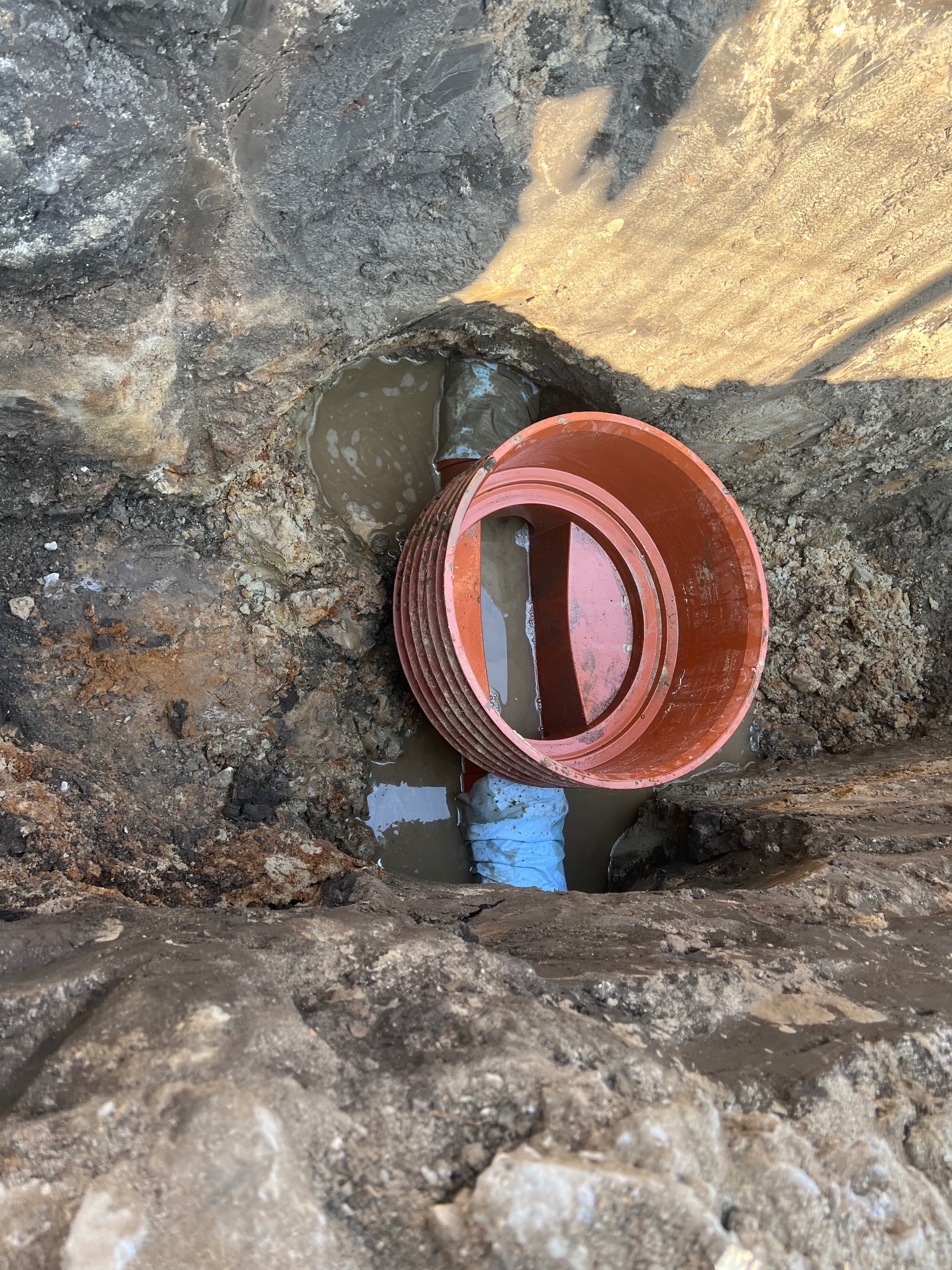 Wykonany wykop z pomarańczową rurą kanalizacyjną w trakcie montażu, widoczne połączenie z niebieską rurą, w wykopie stoi woda.