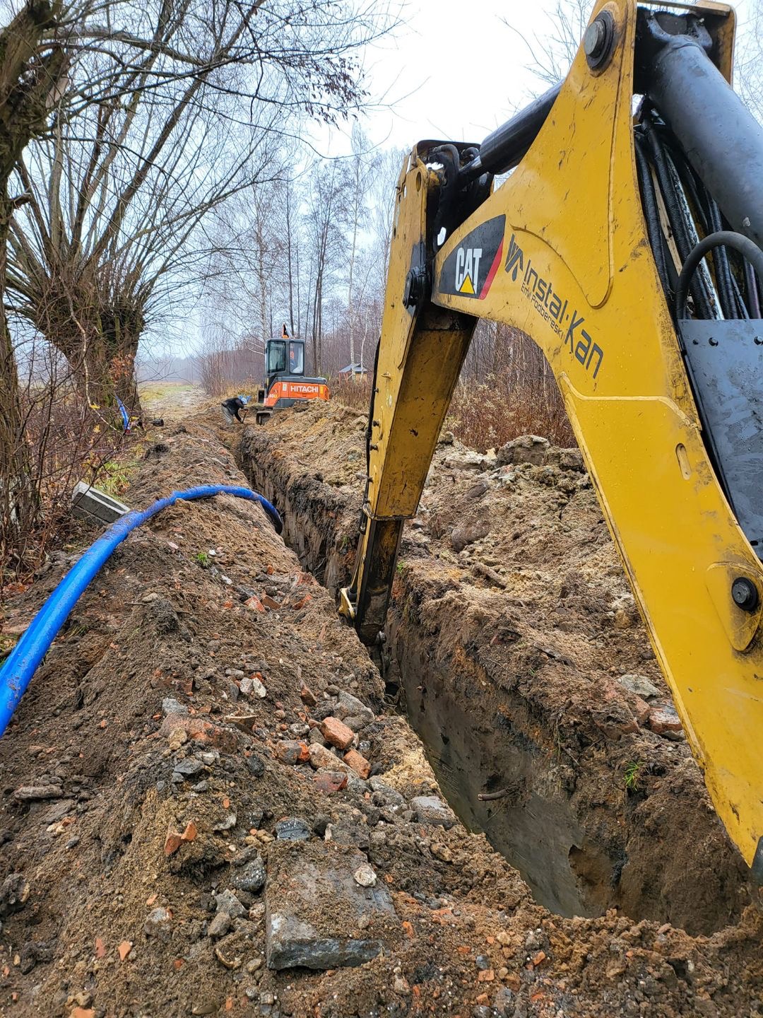 Żółty koparka CAT kopie głęboki wykop w ziemi, widoczny niebieski rurociąg, w tle druga koparka Hitachi, teren budowy w pochmurny dzień.