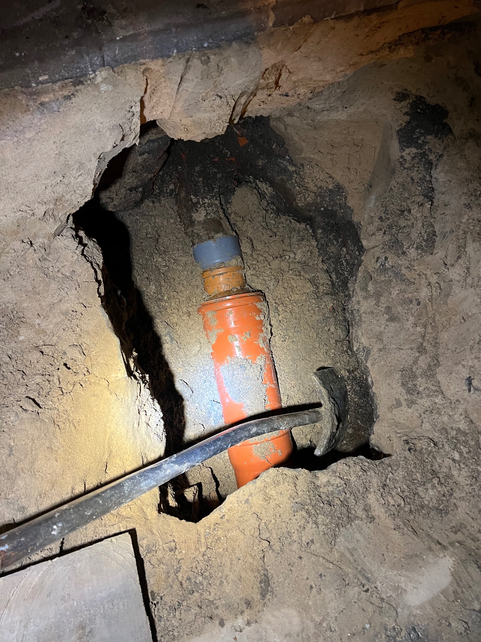 Pomarańczowa rura kanalizacyjna z szarą złączką, częściowo zakopana w ziemi, oświetlona od góry, z widocznym narzędziem hydraulicznym w tle.