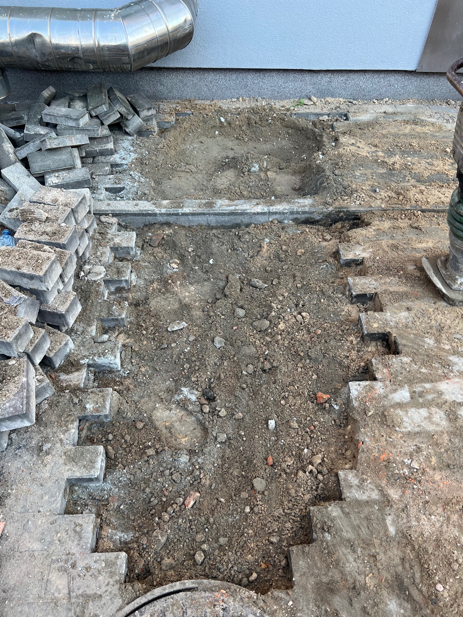 Przygotowanie podłoża pod kostkę brukową: wykop z częściowo ułożonymi krawężnikami i stosem kostki obok metalowej rury wentylacyjnej