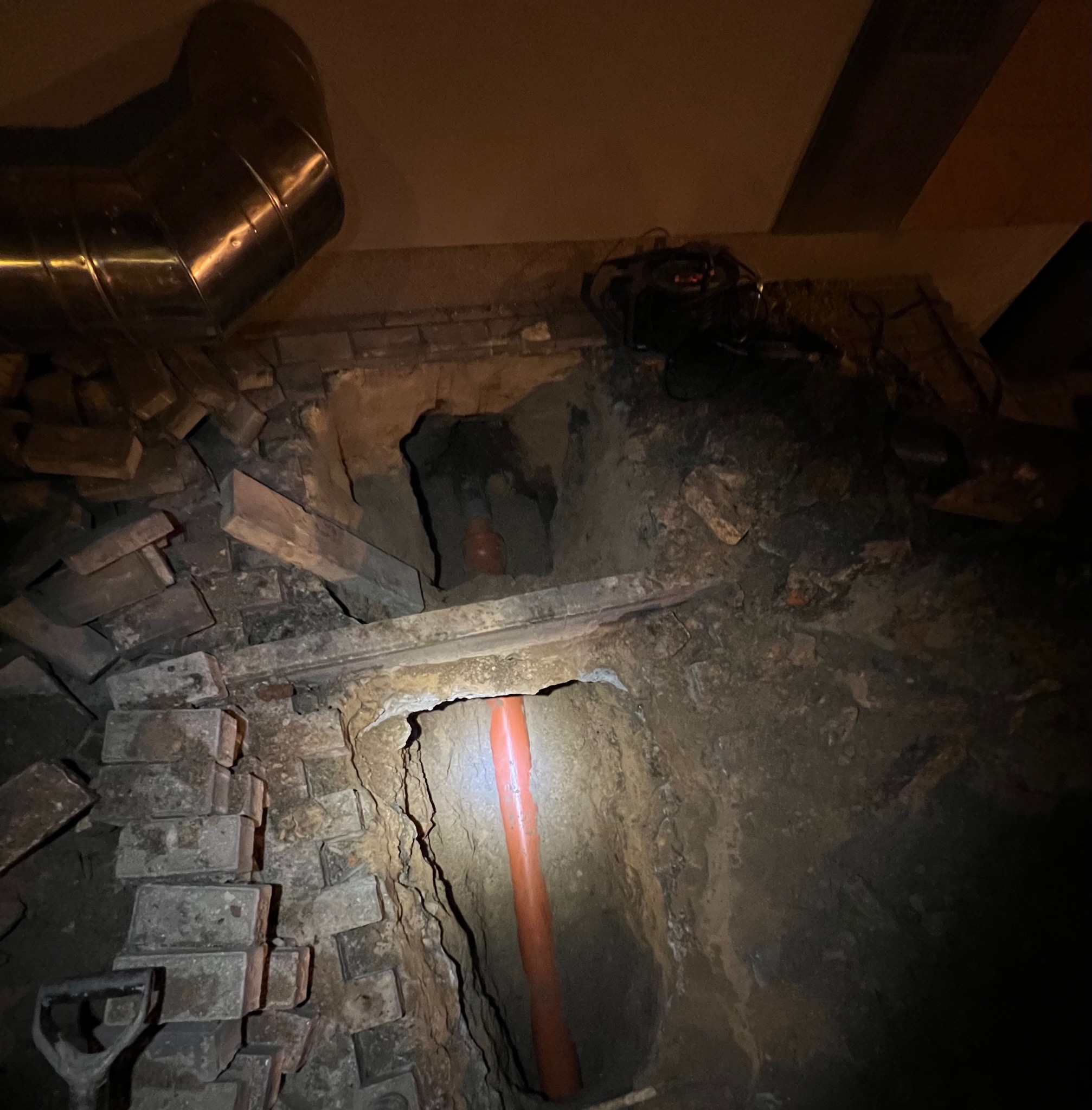 Wykop z widocznymi fragmentami rur kanalizacyjnych, cegłami i elementami instalacji wentylacyjnej w słabym oświetleniu.