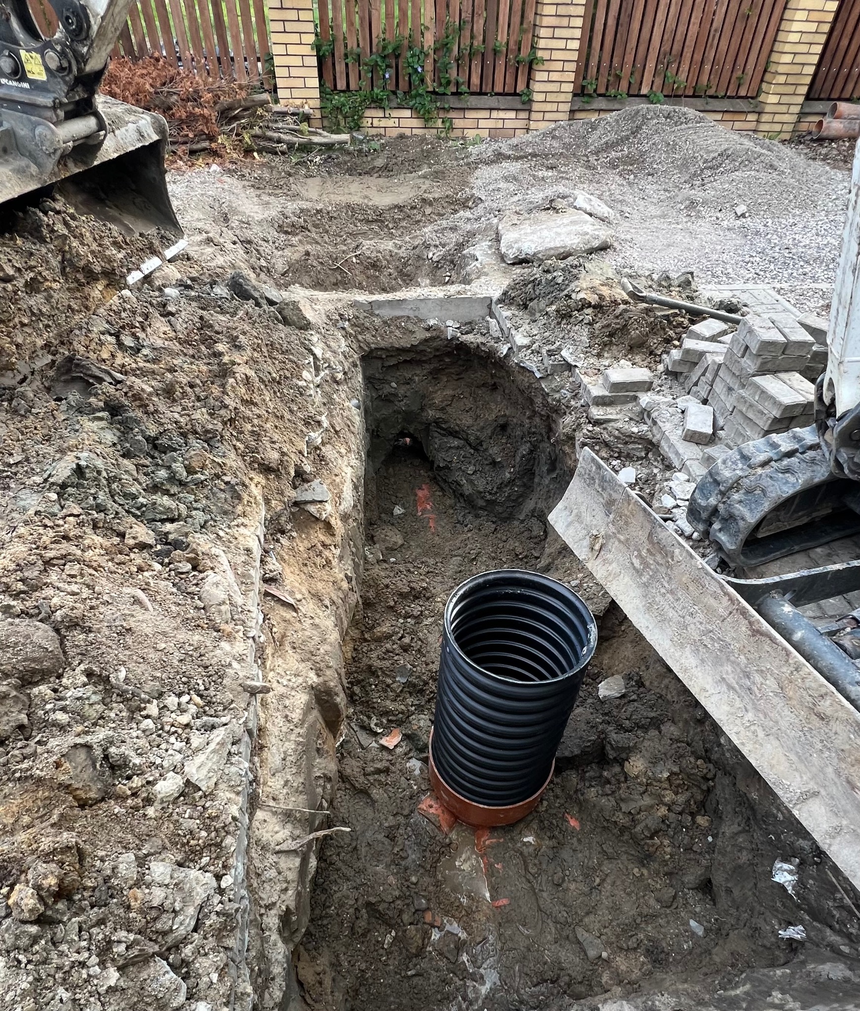 Wykop pod instalację z widoczną karbowaną rurą kanalizacyjną, obok sterta kostki brukowej i fragment koparki.
