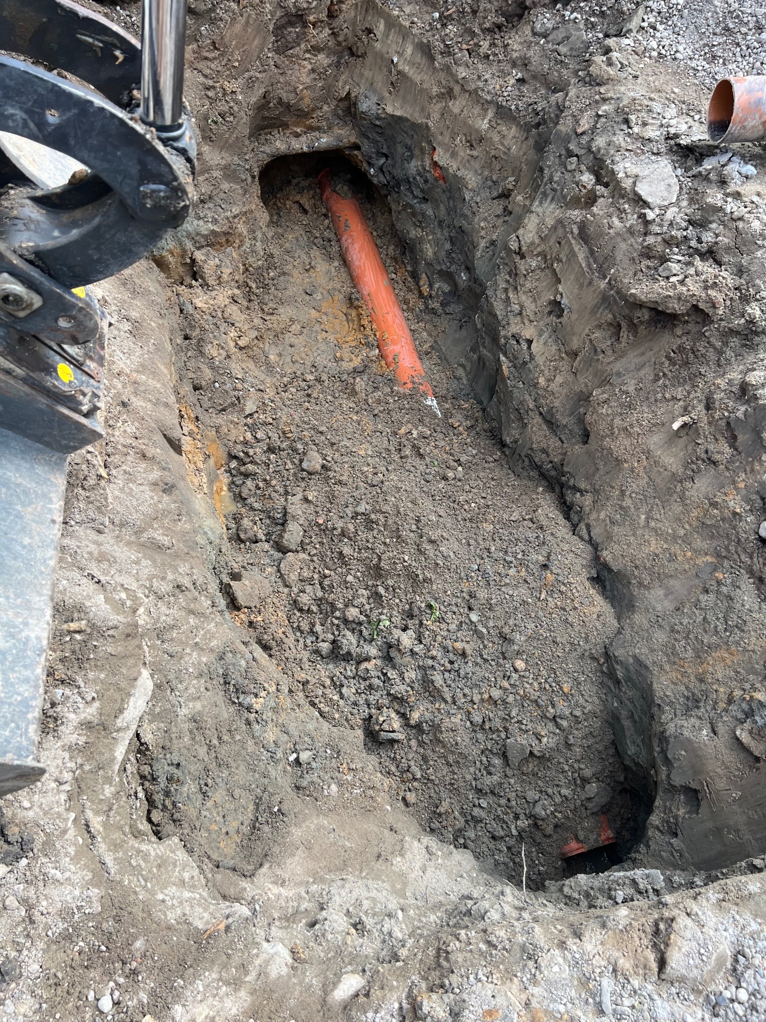 Wykop w ziemi z widocznymi pomarańczowymi rurami, częściowo zasypany gruzem i ziemią, obok ramię koparki.