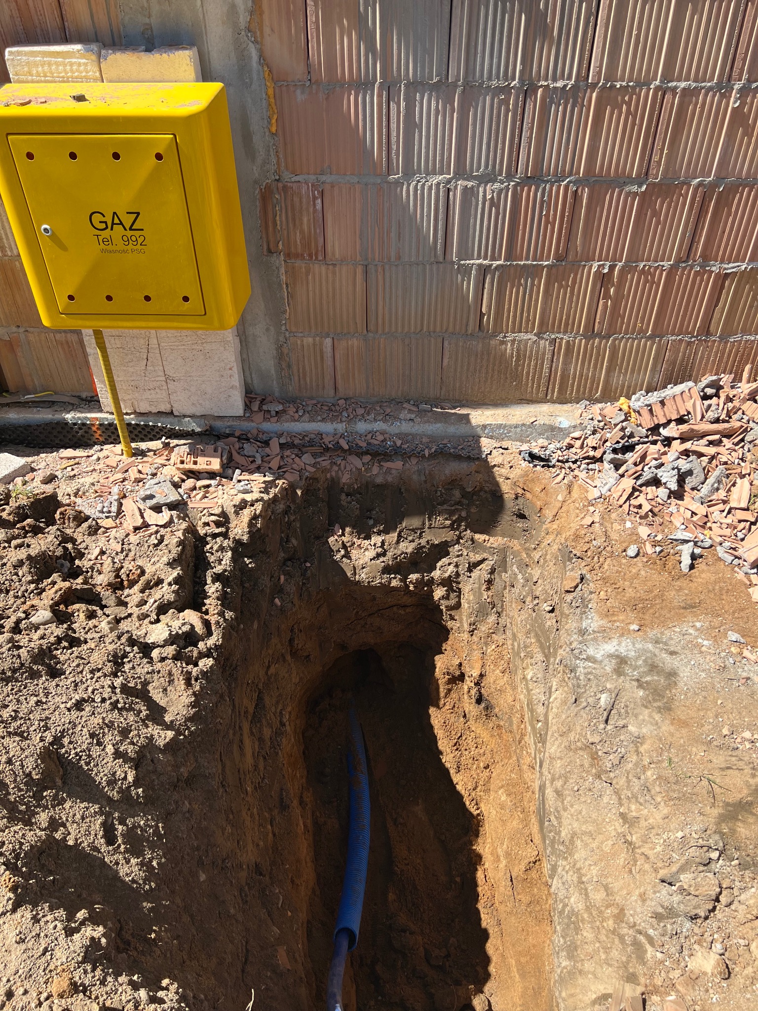 Wykonany wykop w ziemi, widoczna niebieska rura wewnątrz, obok ściana z cegły kratówki i żółta skrzynka gazowa z napisem 'GAZ Tel. 992'.