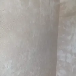 Teksturowana ściana w odcieniach beżu i bieli z widoczną taśmą malarską przy suficie, ukazująca narożnik pomieszczenia i fragment drewnianej podłogi.