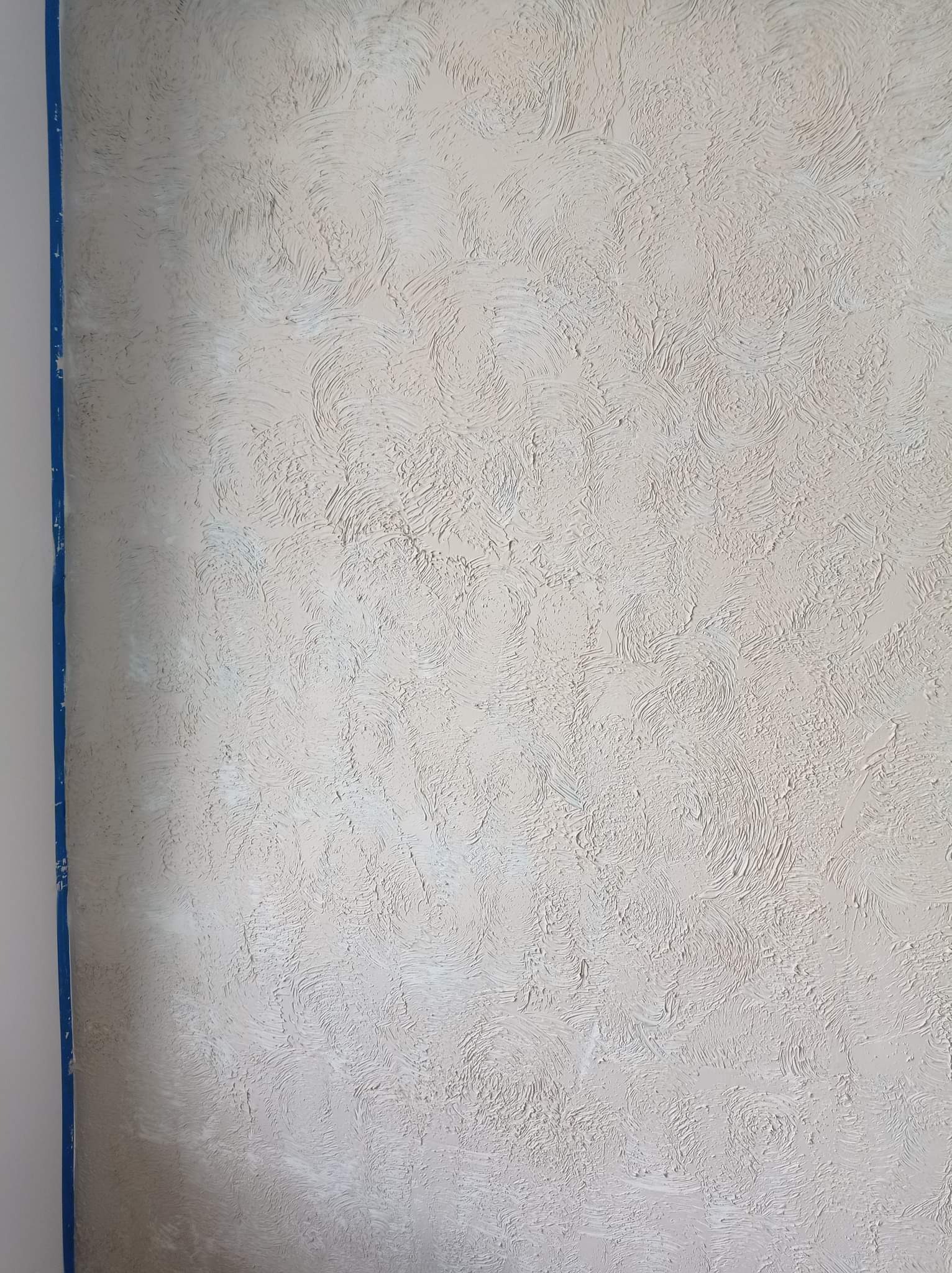 Dekoracyjna struktura gładzi w odcieniach beżu i bieli, wykonana techniką wirującą na ścianie w Kaliszu, zabezpieczona niebieską taśmą malarską przy krawędzi.