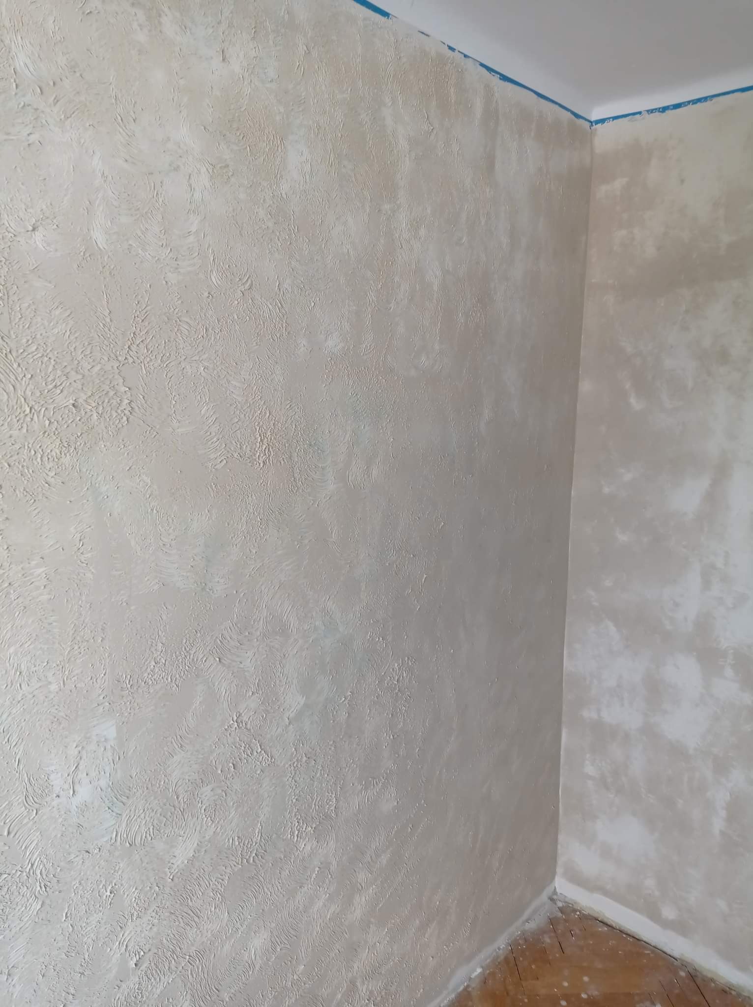 Teksturowana ściana w odcieniach beżu i bieli z widoczną taśmą malarską przy suficie, ukazująca narożnik pomieszczenia i fragment drewnianej podłogi.