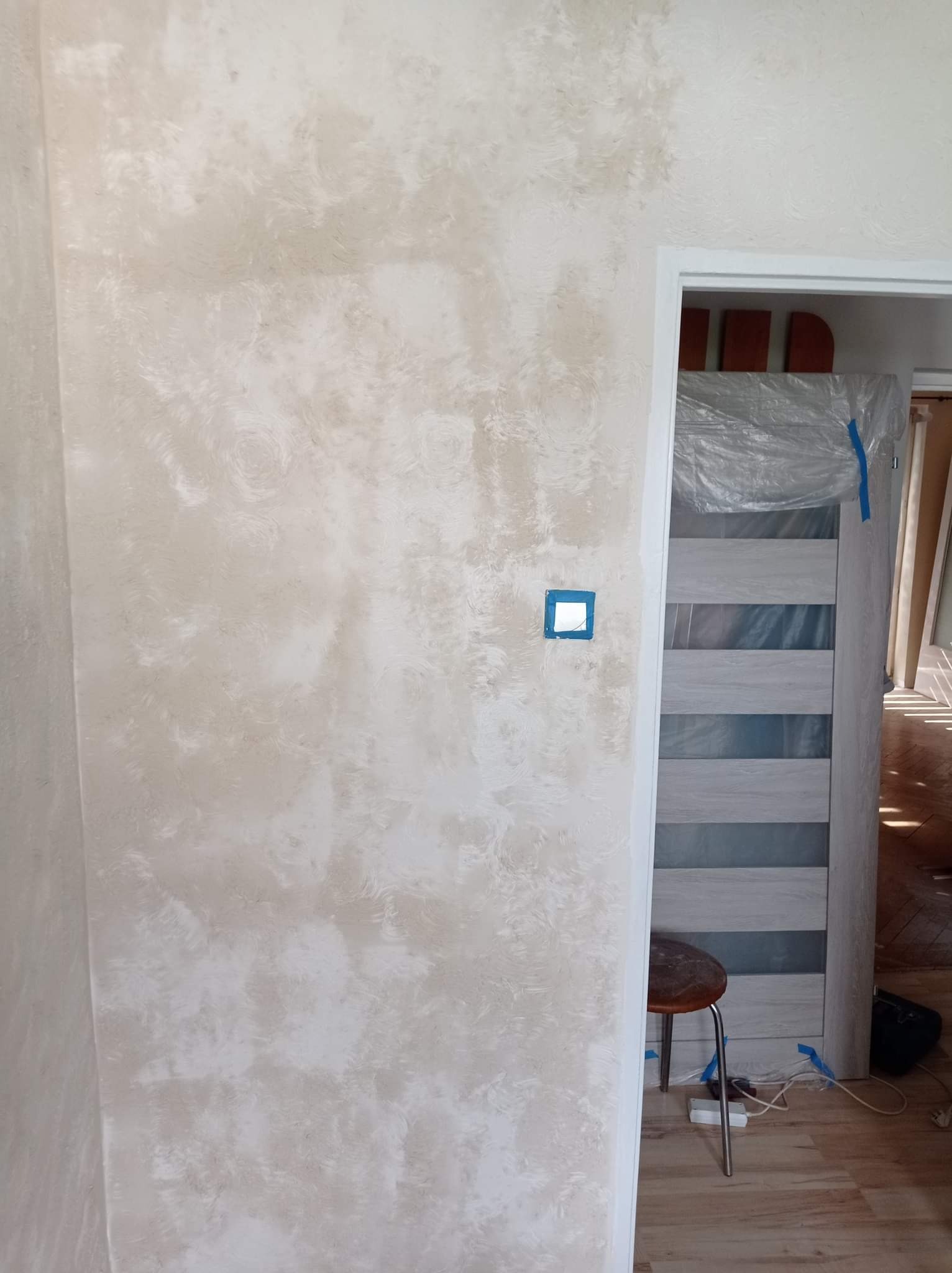 Ściana z dekoracyjną, beżową strukturą, widoczny otwór na włącznik zabezpieczony taśmą, drzwi z okleiną w folii ochronnej, stołek i kable na podłodze.