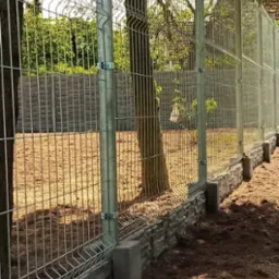 Nowoczesne ogrodzenie panelowe z metalowej siatki zamontowane na betonowej podmurówce, widoczne słupki mocujące i drzewa w tle.