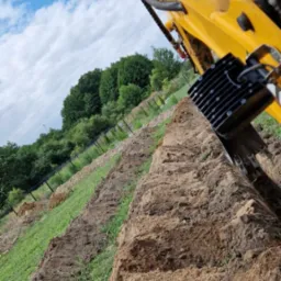 Żółta koparka podczas tworzenia wykopu w ziemi, widok z perspektywy operatora, niebo z chmurami w tle.