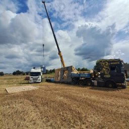 Kujawskie Prefaby - Montaż prefabrykowanej ściany domu na fundamencie za pomocą dźwigu, widoczny transport na lawecie i ciężarówka z logo firmy na tle pola i zachmurzonego nieba.