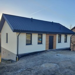 Kujawskie Prefaby - Gotowy dom z ciemną blachodachówką i jasną elewacją, z drewnianymi akcentami przy drzwiach wejściowych. Widoczne czarne okna i rynny. Teren wokół domu jeszcze niezagospodarowany.