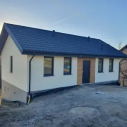 Gotowy dom z ciemną blachodachówką i jasną elewacją, z drewnianymi akcentami przy drzwiach wejściowych. Widoczne czarne okna i rynny. Teren wokół domu jeszcze niezagospodarowany.