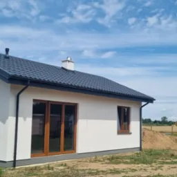 Nowoczesny, parterowy budynek z ciemnym dachem i jasną elewacją, drewniane okna, na działce z widocznym kopcem ziemi. Błękitne niebo z chmurami w tle.