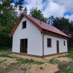 Biały dom z czerwonym dachem na tle lasu i błękitnego nieba. Widoczne brązowe okna i drzwi. Dom otoczony ziemią i zielenią.