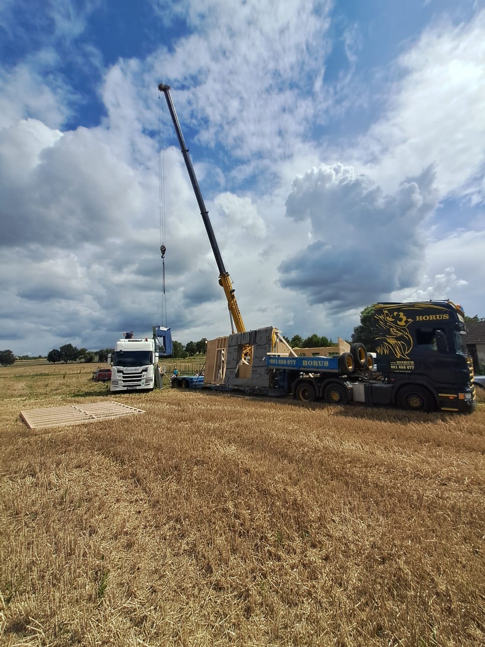 Montaż prefabrykowanej ściany domu na fundamencie za pomocą dźwigu, widoczny transport na lawecie i ciężarówka z logo firmy na tle pola i zachmurzonego nieba.